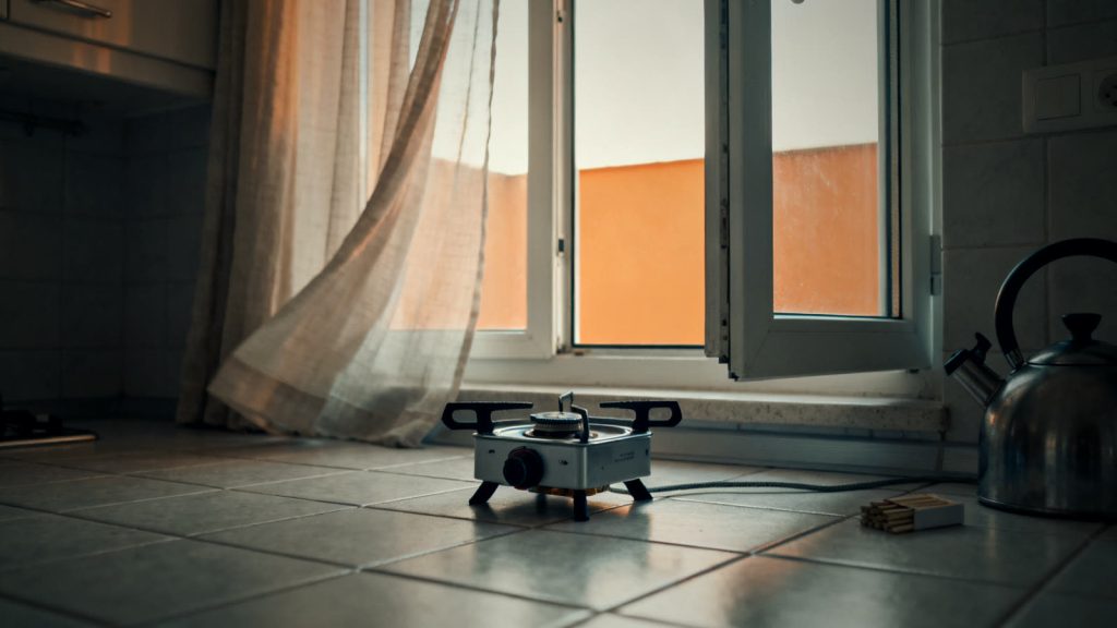 Cocina de gas sobre un suelo de baldosas junto a una ventana abierta, indicación para calentarse y cocinar de forma segura sin electricidad