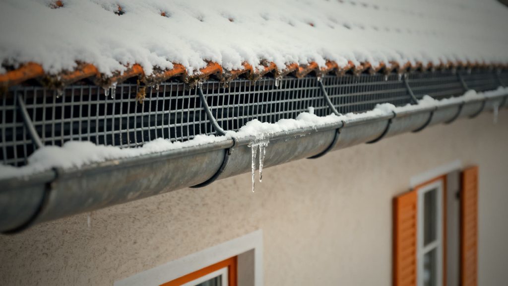 Schneefanggitter am Dachrand mit Schnee und Eiszapfen, wichtig beim Haus winterfest machen